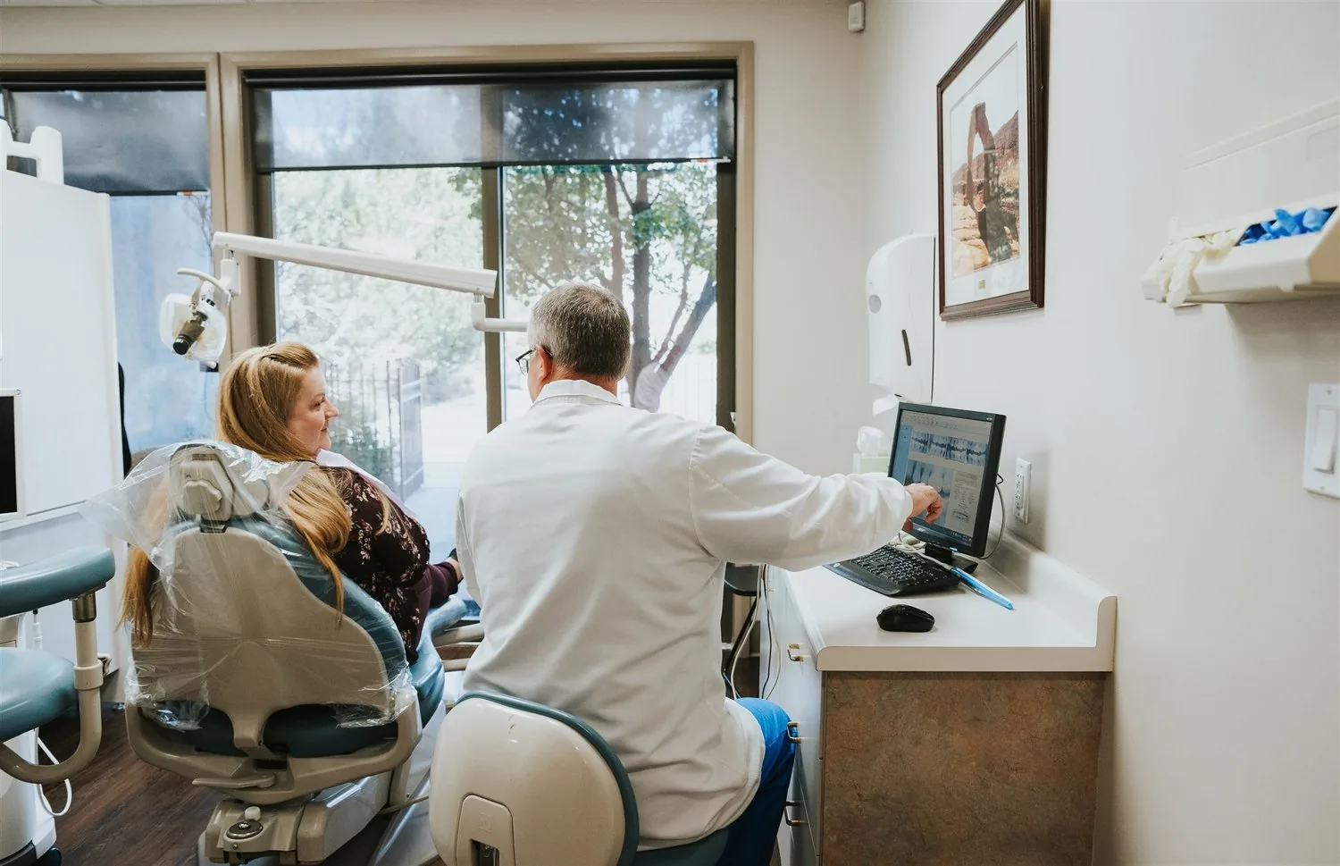 A dentist points to a computer screen while consulting with a patient seated in a dental chair.