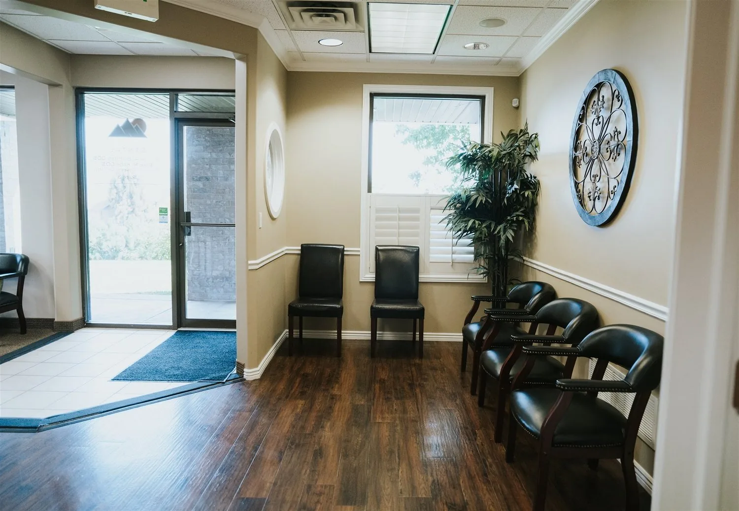 Waiting room with black leather chairs, hardwood flooring, a potted plant, and a glass entryway.